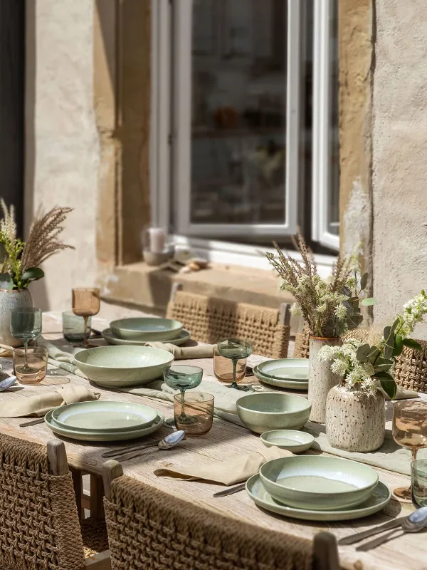 A summery dining table with green plates from the Perlemor collection, glasses and cutlery on a wooden surface. It is framed by wicker chairs and the centrepiece is adorned with decorative vases full of flowers.