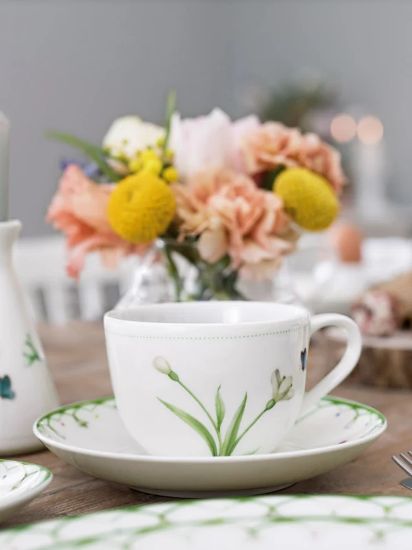 A neatly laid breakfast table with elegant Colourful Spring crockery, a lit candle and a bouquet of fresh flowers.