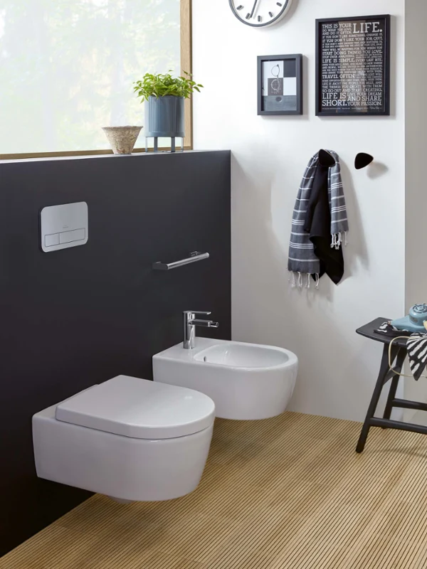 White toilet and bidet from the Avento collection in a modern bathroom with daylight Modern bathroom with a wall-mounted toilet and a bidet from the Avento collection, a towel on a hook, a small plant on a shelf and a clock. A window provides natural light.
