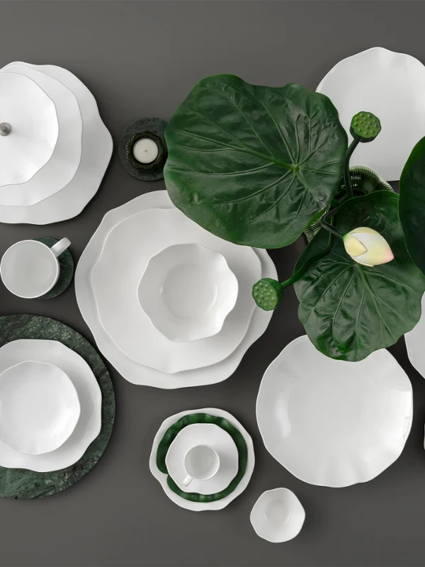 Weiße Geschirrteile der Kollektion Pura auf einer dunklen Fläche stehen. Weiße Geschirrteile der Kollektion Pura auf einer dunklen Fläche mit großen grünen Blättern und einer einzelnen weißen Blume als Dekoration.