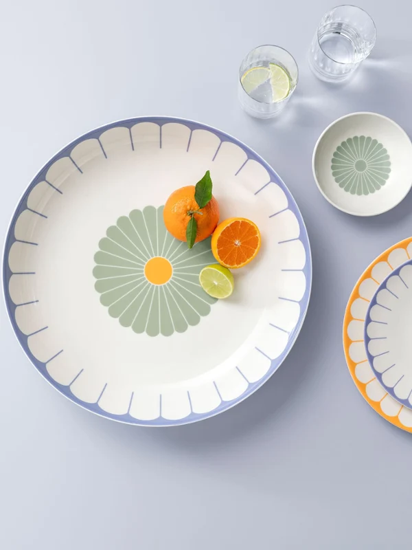 A large plate with orange slices and a mint leaf, next to two smaller plates and two glasses of water, on a light grey surface.