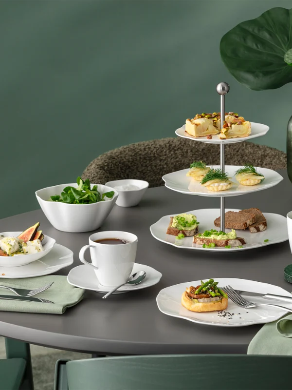 Ein runder Tisch mit weißem Geschirr der Kollektion Pura vor einem grünen Hintergrund Ein runder Tisch mit weißem Geschirr der Kollektion Pura, verschiedenen Backwaren, Salat, Tee und grünem Blattdekor vor einem grünen Hintergrund.