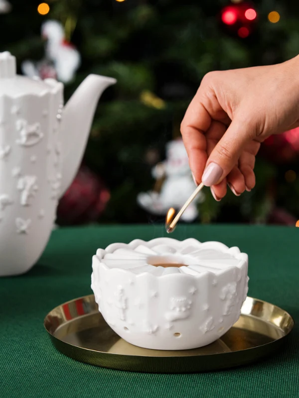 Toy's Delight Royal Classic warmer A hand lights a candle in a white ceramic holder, from Villeroy & Boch, on a festively set table with a teapot, cookies, and a wrapped gift.