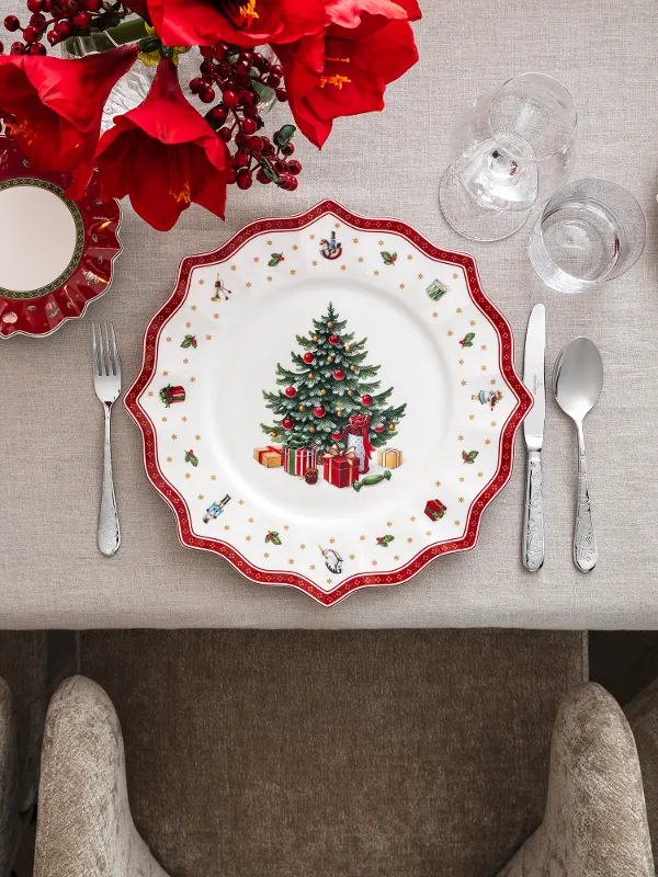 Decorazioni per la tavola festose con piatti con motivi di alberi di Natale, circondati da posate, bicchieri e una composizione di fiori rossi su una tovaglia beige.