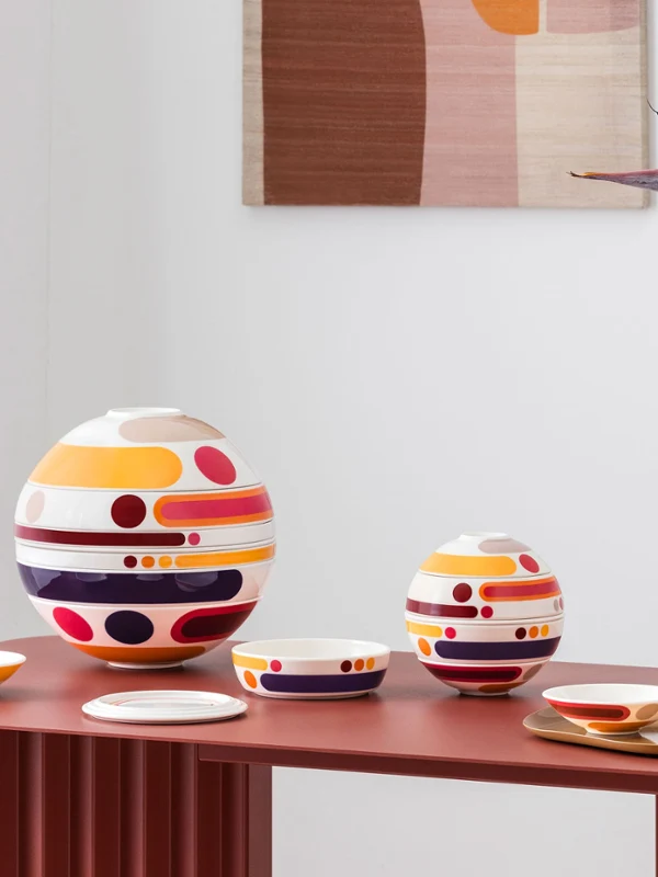 La petite boule miami and la boule miami arranged on a red table Colorful, multi-piece tableware sets from la petite boule miami, circular design objects in large and small sizes, stand as decoration next to a vase with abstract patterns and orange flowers on a red table.