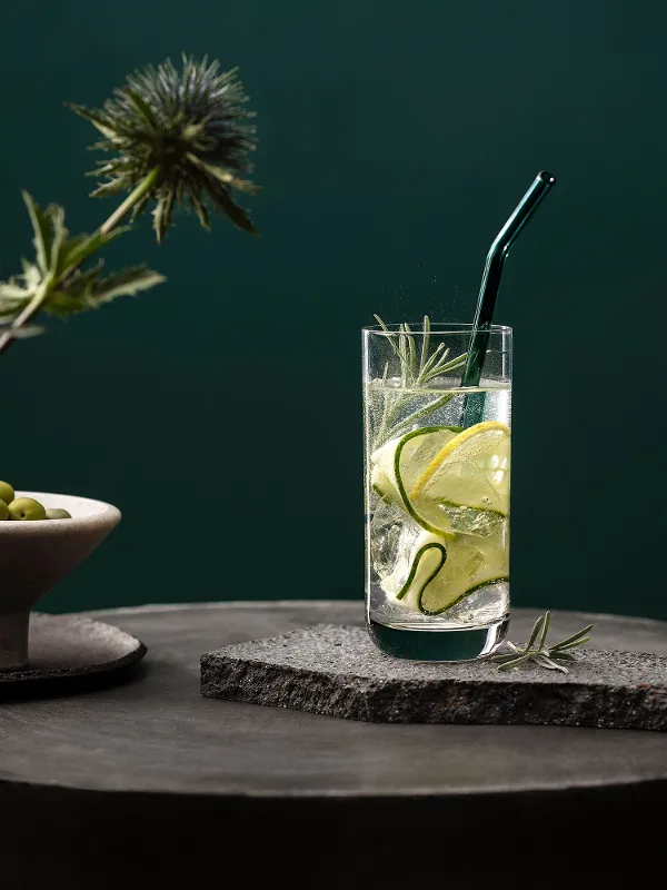 A La Divina glass with cucumber slices and rosemary on a stone surface with a green background. An Artesano glass straw is in the drink and next to it is a bowl of olives.