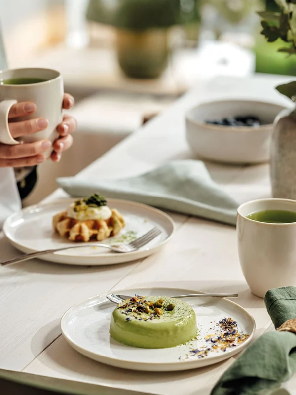 Nahaufnahme eines natürlich dekorierten Esstisches mit Villeroy & Boch Crafted Cotton Porzellan, Piemont Besteck, einer beigen Vase und grünen Servietten, mit einer Frau, die am Tisch sitzt.