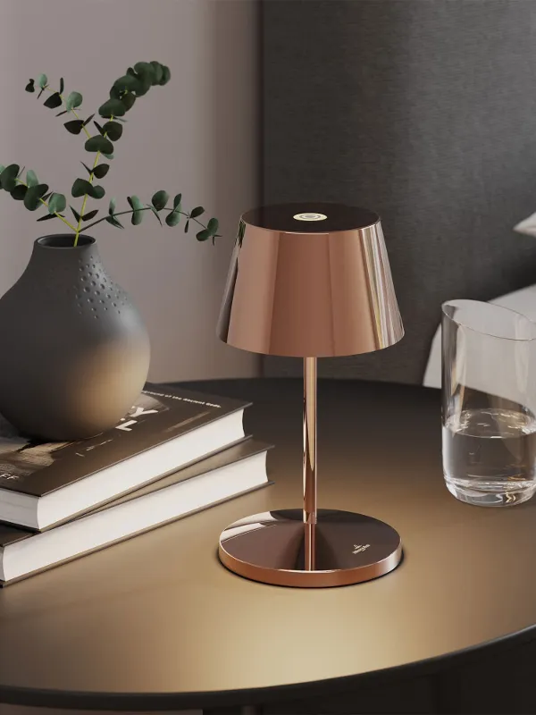 Runder Nachttisch mit einer goldenen Lampe, zwei Büchern, einer dunklen Vase mit Grünpflanzen und einem Glas Wasser neben einem Bett.
