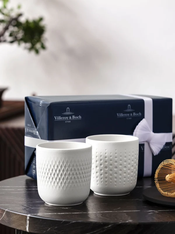 Dwa białe kubki collier przed drzewkiem bonsai Dwie białe filiżanki na stole z trzepaczką do herbaty i pudełkiem prezentowym, przed drzewkiem bonsai.