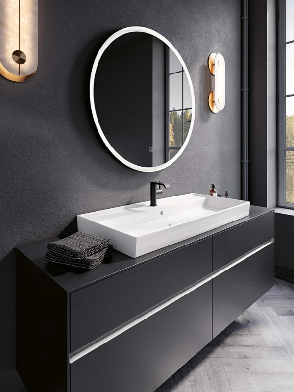 Modern bathroom with a white Collaro washbasin and Subway 3.0 bathroom furniture from Villeroy & Boch. Modern bathroom with an elegant black washstand, a white Collaro washbasin from Villeroy & Boch, a round backlit mirror and wall-mounted lights. A large window provides natural light and an outside view.