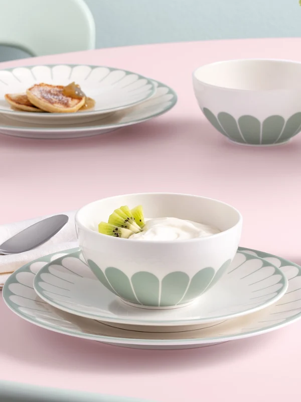 A table set with plates, bowls, and mugs featuring a flower pattern, with food and a Fleur glass vase on a pink tablecloth.