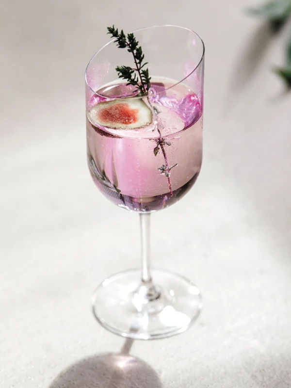 Een roze cocktail, gegarneerd met rozemarijn en fruit, wordt elegant gepresenteerd in een Villeroy & Boch NewMoon wijnglas op een witte achtergrond.
