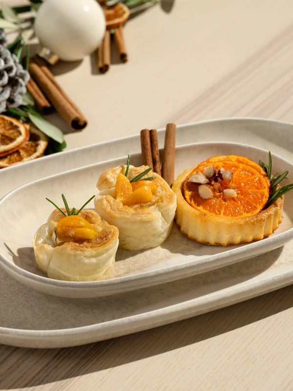 Tres pasteles variados en un plato ovalado blanco Perlemor, con rodajas de naranja secas y ramas de canela de fondo.