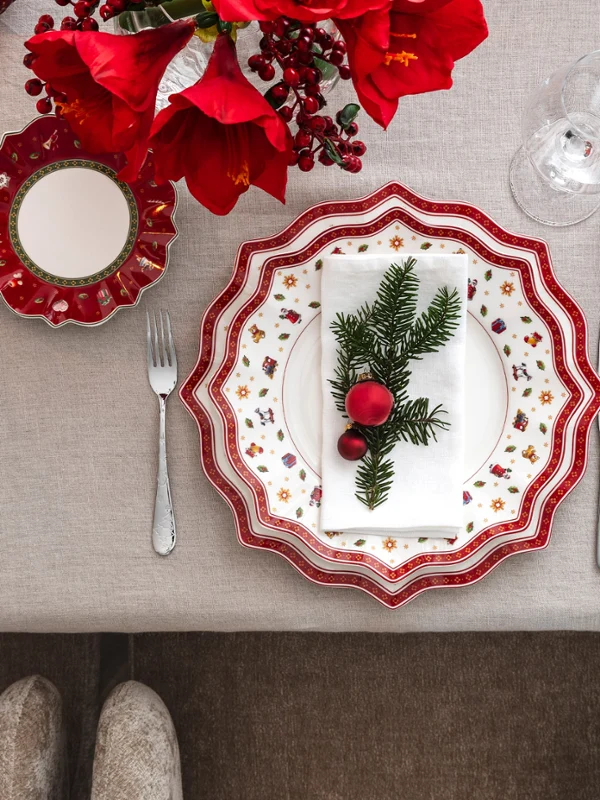 Une vue de dessus d’une élégante table de fête avec la collection Toy’s Delight de Villeroy & Boch, avec des assiettes rouges et blanches, des couverts de table argentés et une serviette de fête décorée de pin et d’ornements.