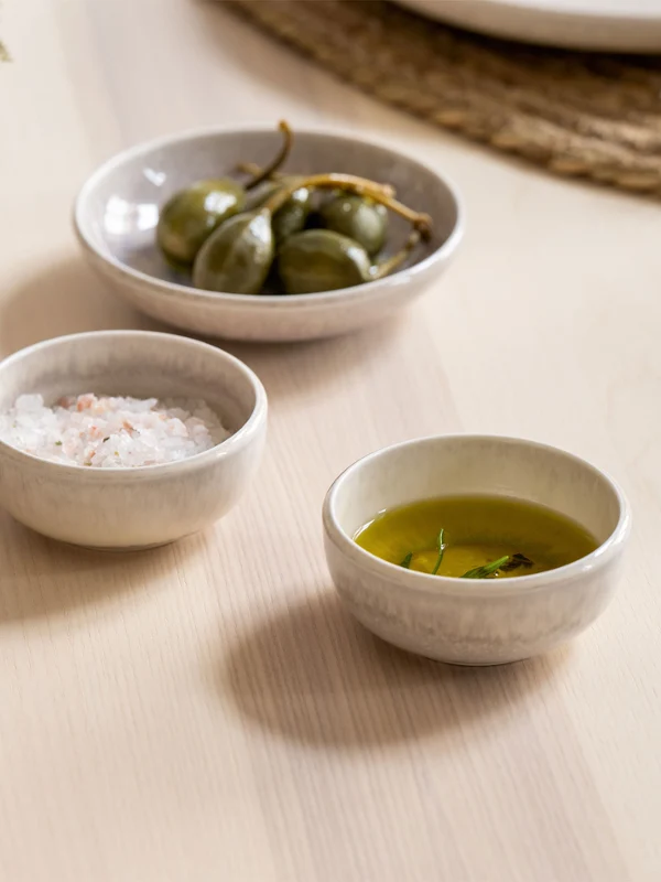 There are three small mother-of-pearl dip bowls on a table. On a table are three small Perlemor dip bowls containing salt, green olives and olive oil.