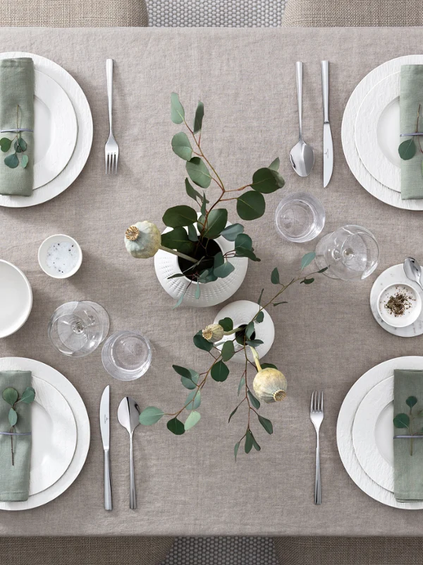 Modern dining table for six people with white Manufacture plates, cutlery and green floral decoration, seen from above.