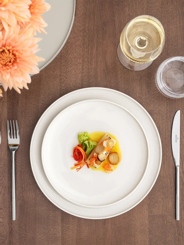 Ein formeller gedeckter Tisch der Afina Kollektion  mit einem kleinen Teller, einem Weinglas, einem Wasserglas, Besteck, einer Serviette und einer Blumenvase.
