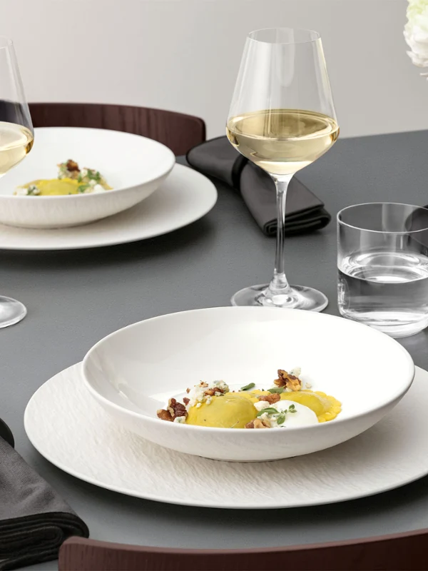 A formal table setting with a white bowl on a white plate, glassware, and silver cutlery on a grey table and food.