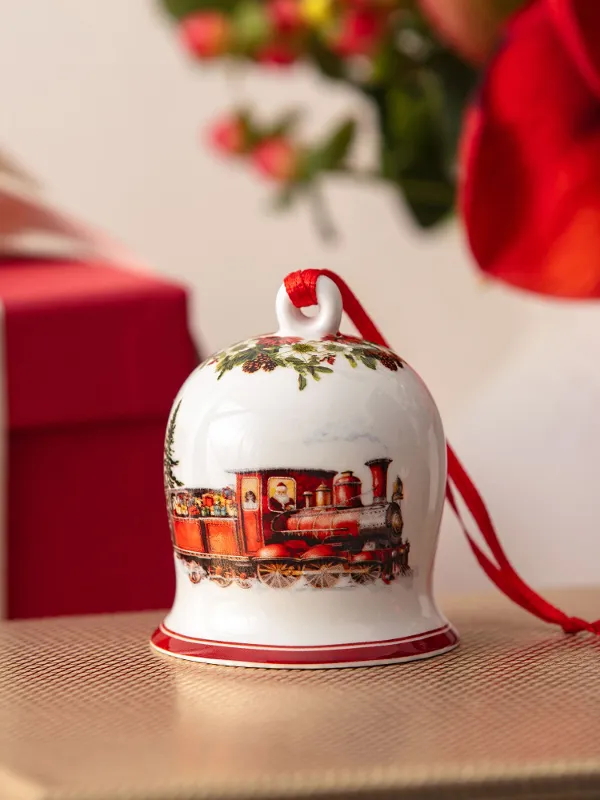 A ceramic bell with a train motif sits on a golden box, with a red gift and flowers in the background.