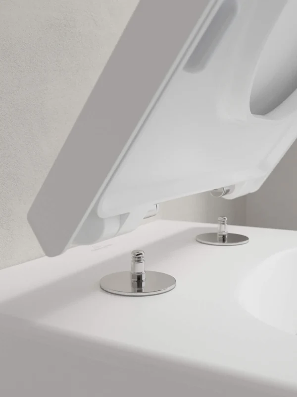 Venticello toilet seat attached to a toilet Close-up of a white toilet with an open lid showing the fixing screws on the toilet seat. The background is a light-grey wall.