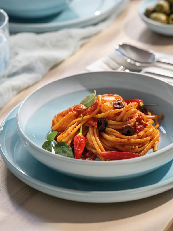 A pasta bowl stands on a blue Perlemor aqua plate. A pasta bowl from the Perlemor Aqua collection stands on a blue Perlemor Aqua plate with spaghetti, tomato sauce, and vegetables, next to cutlery and a salad.