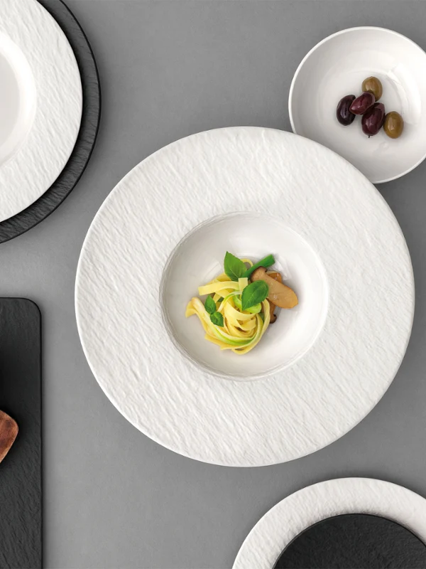 A formal table setting with a white past plate with food in it on a grey table.