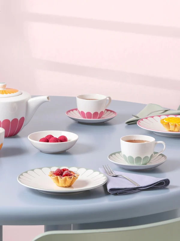 A tea set with colorful Fleur tableware on a light blue round table A pastel-colored tea set with cups, saucers, plates with colorful floral patterns from the Fleur collection, and small desserts on a light blue round table.