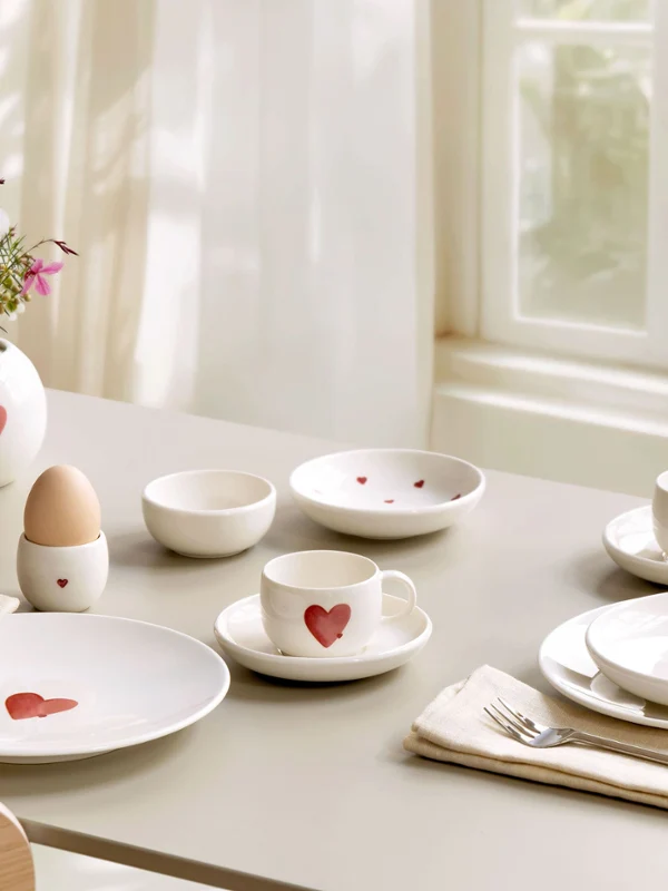Der Frühstückstisch ist mit weißem Geschirr mit roten Herzmotiven von with love, Blumen in Vasen von with love und ordentlich angeordneten Utensilien gedeckt.