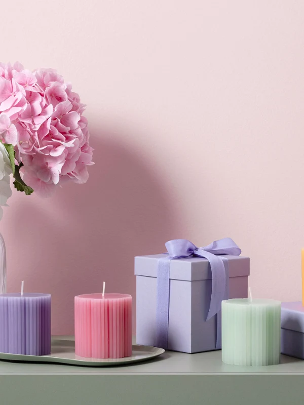 Fleur candles in pastel colors as a gift idea Fleur glass vase with white and pink flowers, pastel-colored Fleur candles in the shape of a cloverleaf, and purple gift boxes on a green surface in front of a pink wall.