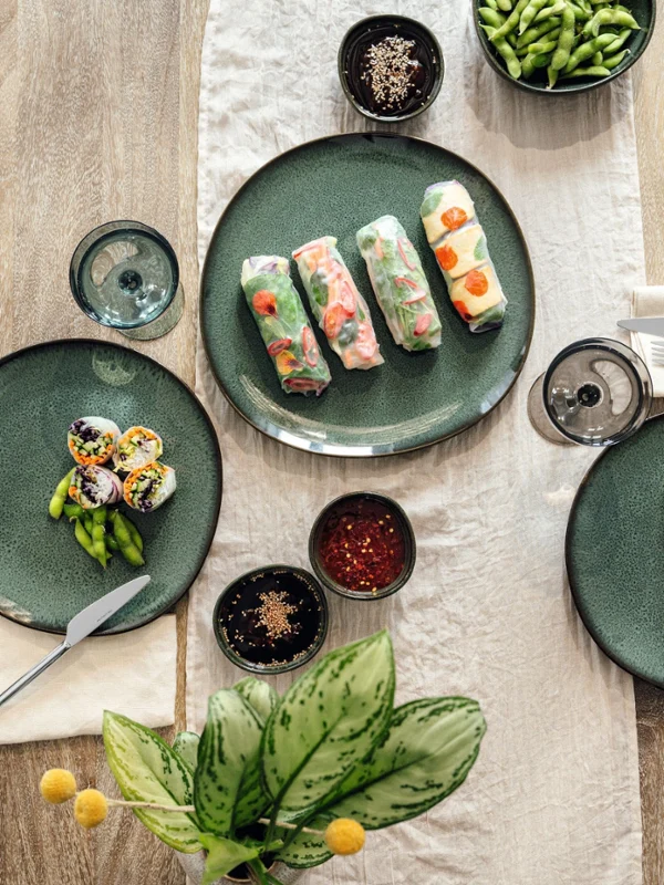 Top view of a wooden table with green crockery, on which sushi rolls, spring rolls, edamame and sauces are laid out.