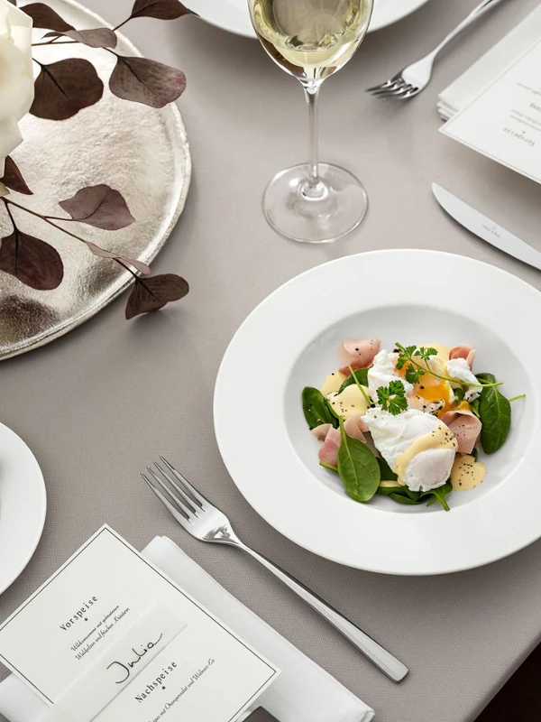 Elegante arreglo de mesa compuesto por un plato Royal de Villeroy & Boch con ensalada, complementado con una copa de vino blanco, una tarjeta de menú y una guarnición de pan asado. Un elegante centro de mesa con rosas blancas.