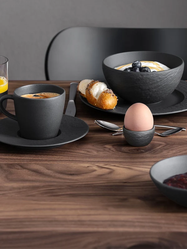 Ein Frühstücksgedeck mit Kaffee, Saft, Croissant, Marmelade und einem weichgekochtem Ei auf einem Holztisch, grüne Blumen sind zu sehen.