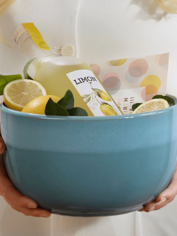 Eine Person hält eine blaue Schale der Perlemor Aqua Kollektion mit einer Flasche Limonade, Zitronenscheiben und grünen Blättern.
