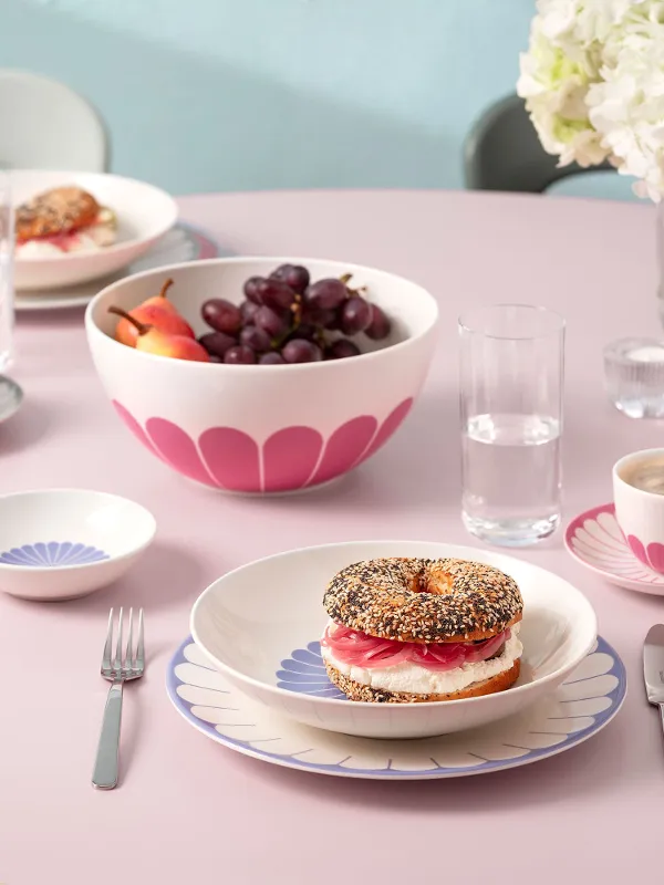 A round pink table with plates full of food, including a filled bagel, a bowl of fruit, cups of coffee and cutlery. In the background is a vase of white flowers.