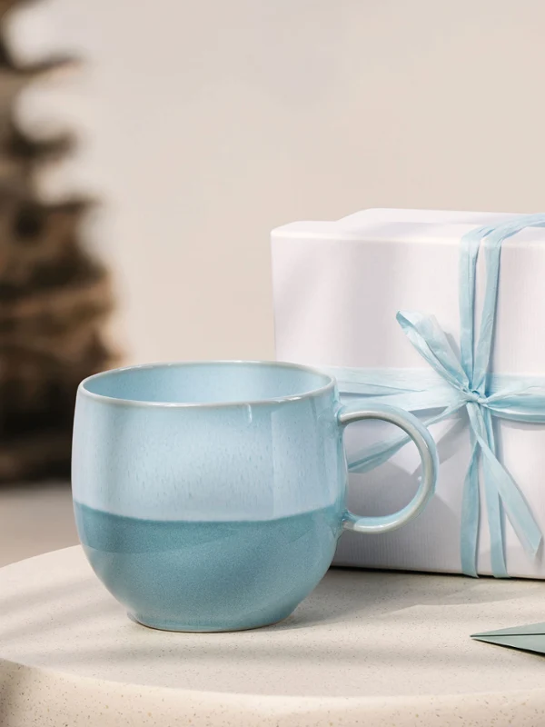 Light blue Perlemor Aqua cup and white gift box with blue ribbon on a round table; blurred background with a plant.