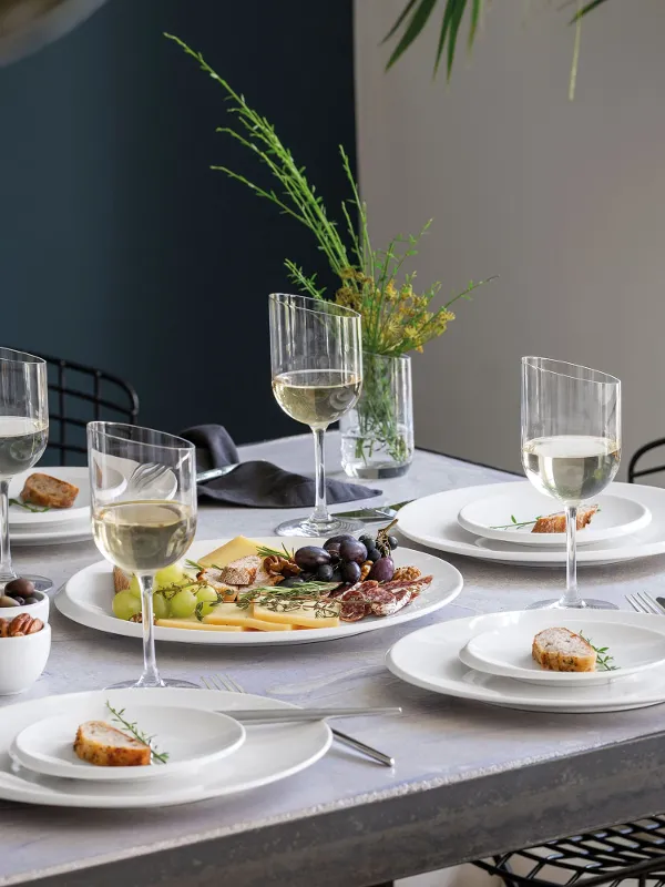 Un tavolo da pranzo con piatti bianchi NewMoon, bicchieri da vino, formaggio, frutta e antipasti, pronto per un pasto.