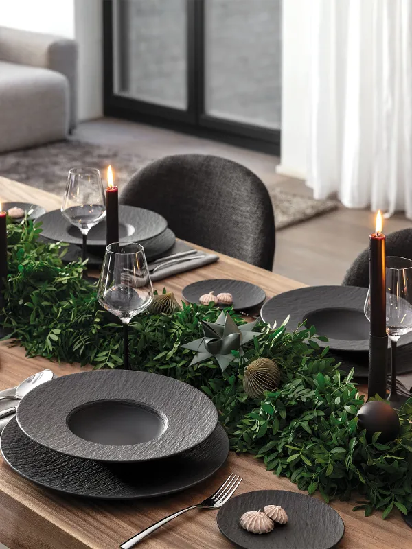 Elegant dining table set with black Manufacture plates, black candles, cutlery and green foliage as a centrepiece in a modern dining room.