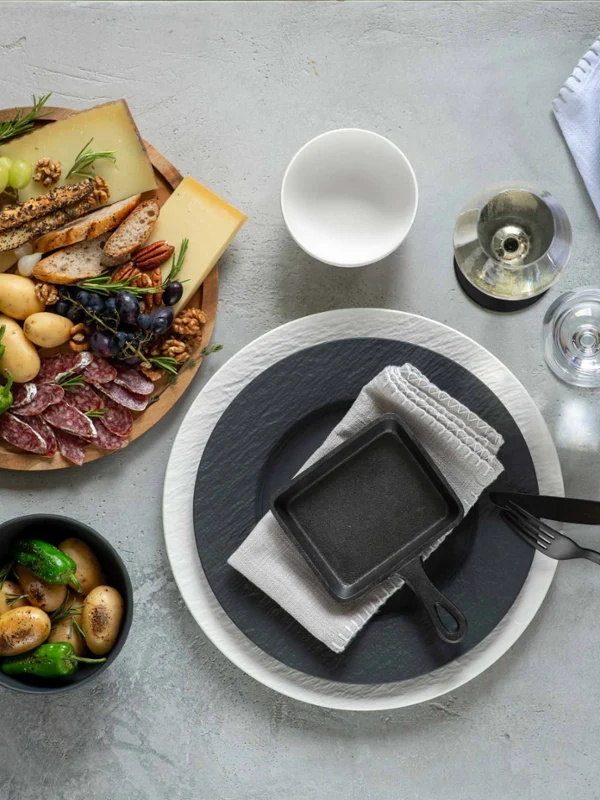 Vista dall'alto di un menu gourmet con tagliere di formaggi e salumi, carciofi, pane, tovagliolo nero con utensili e bevande di accompagnamento su una superficie grigia strutturata ricoperta di stoviglie e posate Manufacture. 