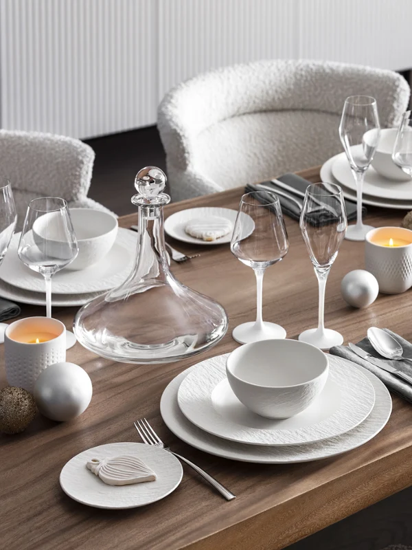 Manufacture white christmas table photographed from the side with candles and flowers A dining table set with Manufacture plates, glassware, silver cutlery, candles, and a white floral centerpiece.