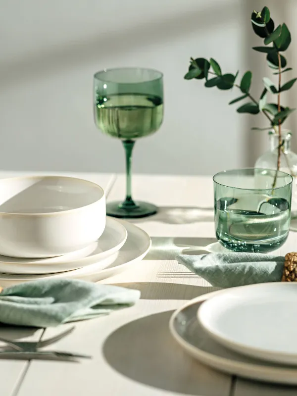 Stół jadalniany z białymi talerzami Crafted, zielonymi kieliszkami like Glass i miskami, a także zielonymi serwetkami ze złotymi uchwytami i dekoracjami z zielonych liści.