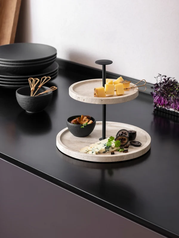 Petites gourmandises sur Travertine de Villeroy & Boch Une étagère en travertin à deux étages avec des cubes de fromage, des raisins, des noix et des herbes sur un plan de travail noir.