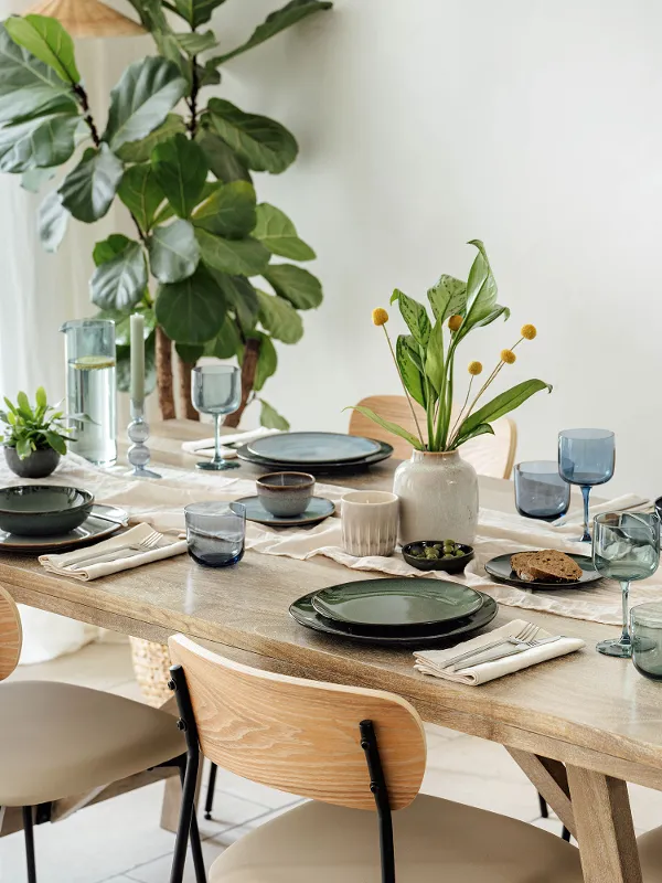 Une table de salle à manger avec des assiettes vertes, des serviettes beiges, des verres bleus et des petites plantes. Des chaises en bois clair entourent la table. Une grande plante est placée près d'une fenêtre.