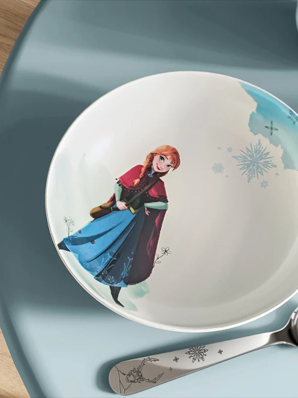 Un cuenco de la colección Disney Frozen está junto a una cuchara sobre una mesa azul claro. Un cuenco de la colección Disney Frozen con un personaje animado está junto a una cuchara sobre una mesa azul claro.