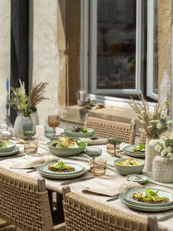 Une table rustique en plein air est dressée avec des assiettes Perlemor remplies de salade, des verres à vin et des centres de table floraux près d'un mur en pierre.