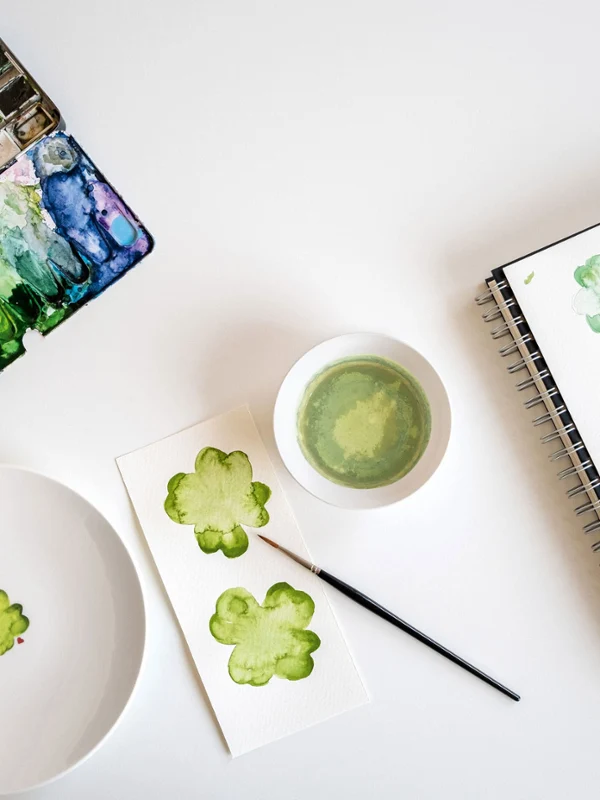 Ein Aquarell-Setup mit einem „With Love“-Teller von Villeroy & Boch und einem Skizzenbuch, geschmückt mit grünen Kleeblatt-Bildern auf einem weißen Tisch.