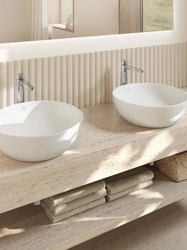 Strefa umywalki w łazience z umywalką w kolorze beżowym Nowoczesna łazienka z dwiema okrągłymi umywalkami Artis na marmurowym blacie, lustrem, przyborami toaletowymi, złożonymi ręcznikami i koszem do przechowywania pod spodem.