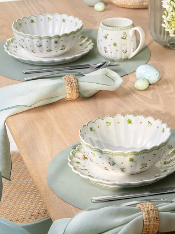A wooden table set with Easter Delight bowls, mugs, plates, napkins, and a small flower arrangement.