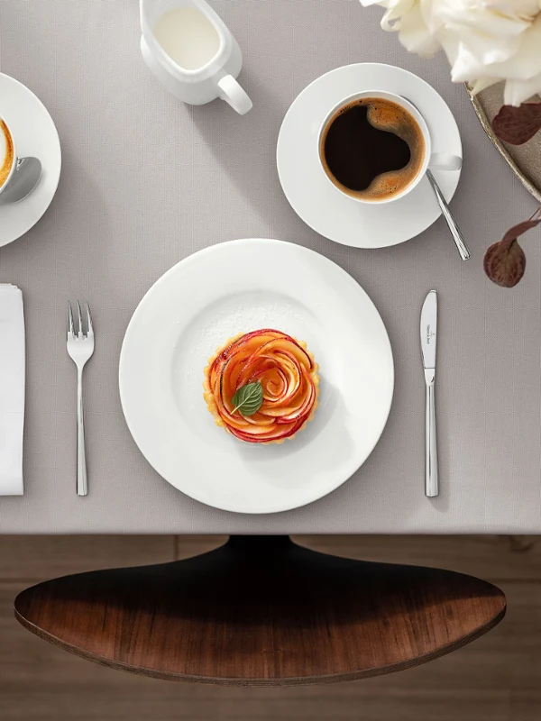 Una bonita mesa de comedor con la colección Royal de Villeroy & Boch, que incluye un plato con comida, una taza de café, un capuchino, cubiertos, servilletas y una jarra de leche. También hay un arreglo floral a la derecha de la mesa.