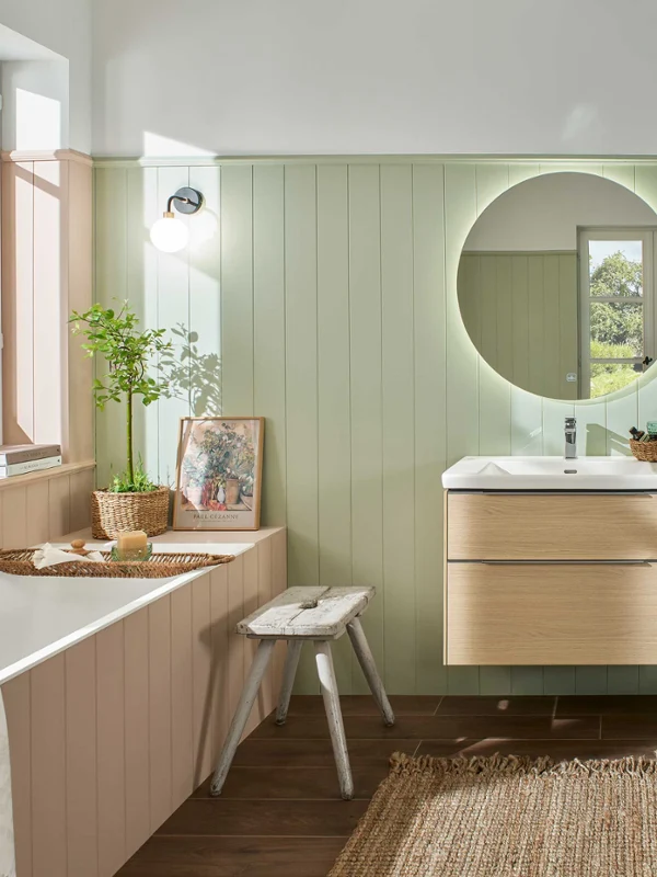 Łazienka z wanną Subway 3.0 marki Villeroy & Boch Łazienka z wanną Subway 3.0 marki Villeroy & Boch, okrągłym lustrem i drewnianą szafką z umywalką. Rośliny i wiklinowe dekoracje nadają jej naturalny charakter. Drewniane panele w pastelowych kolorach podkreślają rustykalną estetykę. Przez okno wpada światło.