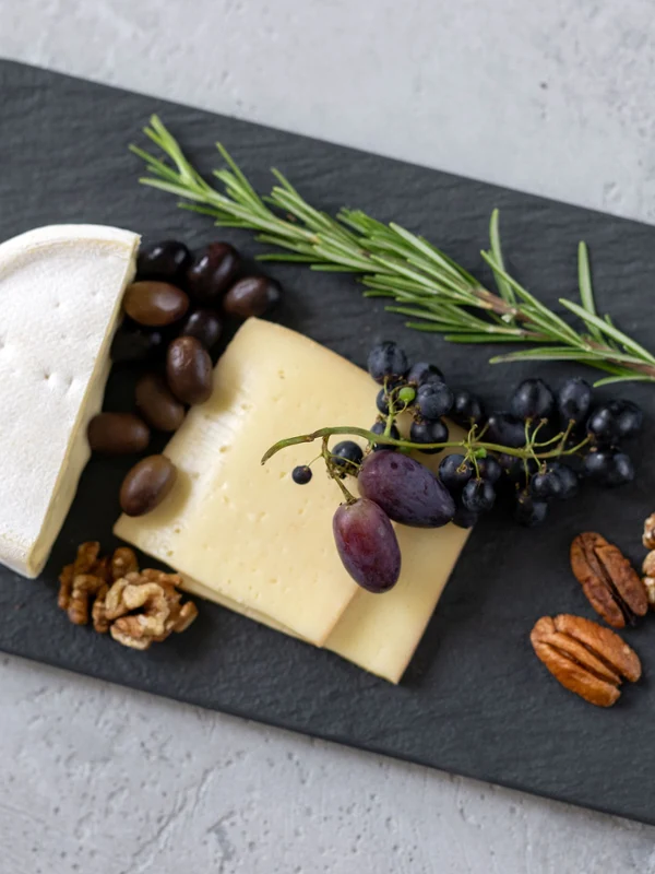 Un plateau à servir Manufacture noir utilisé comme plateau à fromage Un plateau de service carré noir de Manufacture avec des tranches de fromage, un morceau de fromage à pâte molle, des olives, des raisins, des noix, des noix de pécan et une branche de romarin.